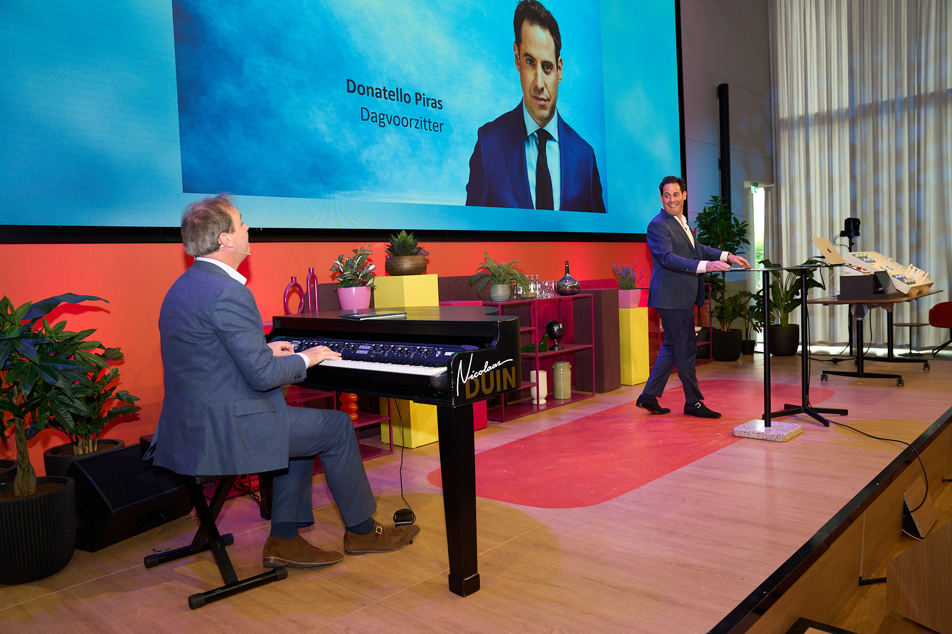 Nicolaas Duin als Muzikaal Spreker achter zijjn piano, en dagvoorzitter Donatello Piras midden op het podium, met daarachter zijn foto op het scherm